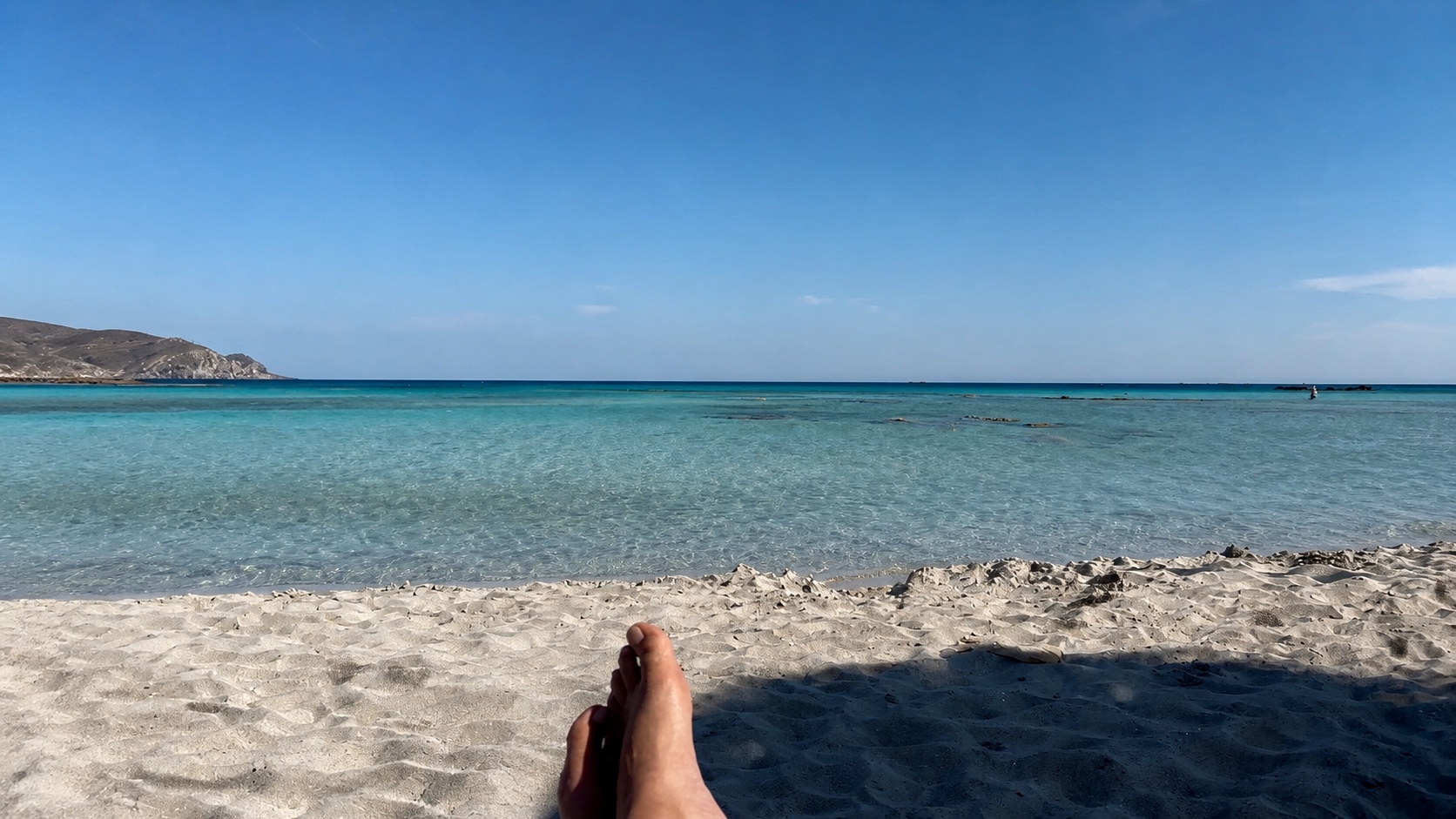 Bare feet rest in shade before the pale sand and turquoise shallows of Elafonisi.