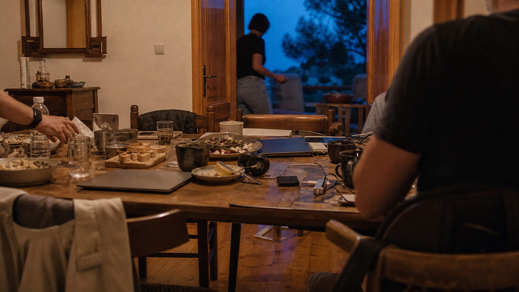 A modest rental dining table fills with food, laptops, cables and anonymous visitors at dusk.